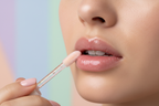 “Close-up of a woman applying lip oil with a doe-foot applicator, soft lighting, glossy lips, pastel background, clean and modern beauty aesthetic.”